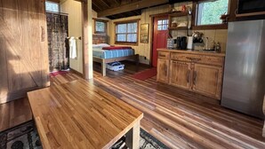 A rustic interior with a wooden floor, a bed with a red headboard, a kitchen with wooden cabinets, and a large window. - Cherry Treesort "Chico's Hideaway" (Rowan County)