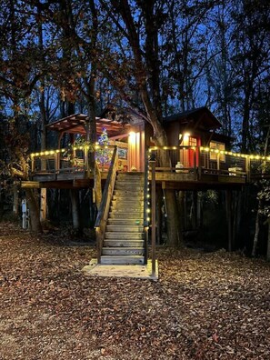 A treehouse with a wooden staircase and a small deck, surrounded by trees and decorated with string lights. - Cherry Treesort "Chico's Hideaway" (Rowan County)