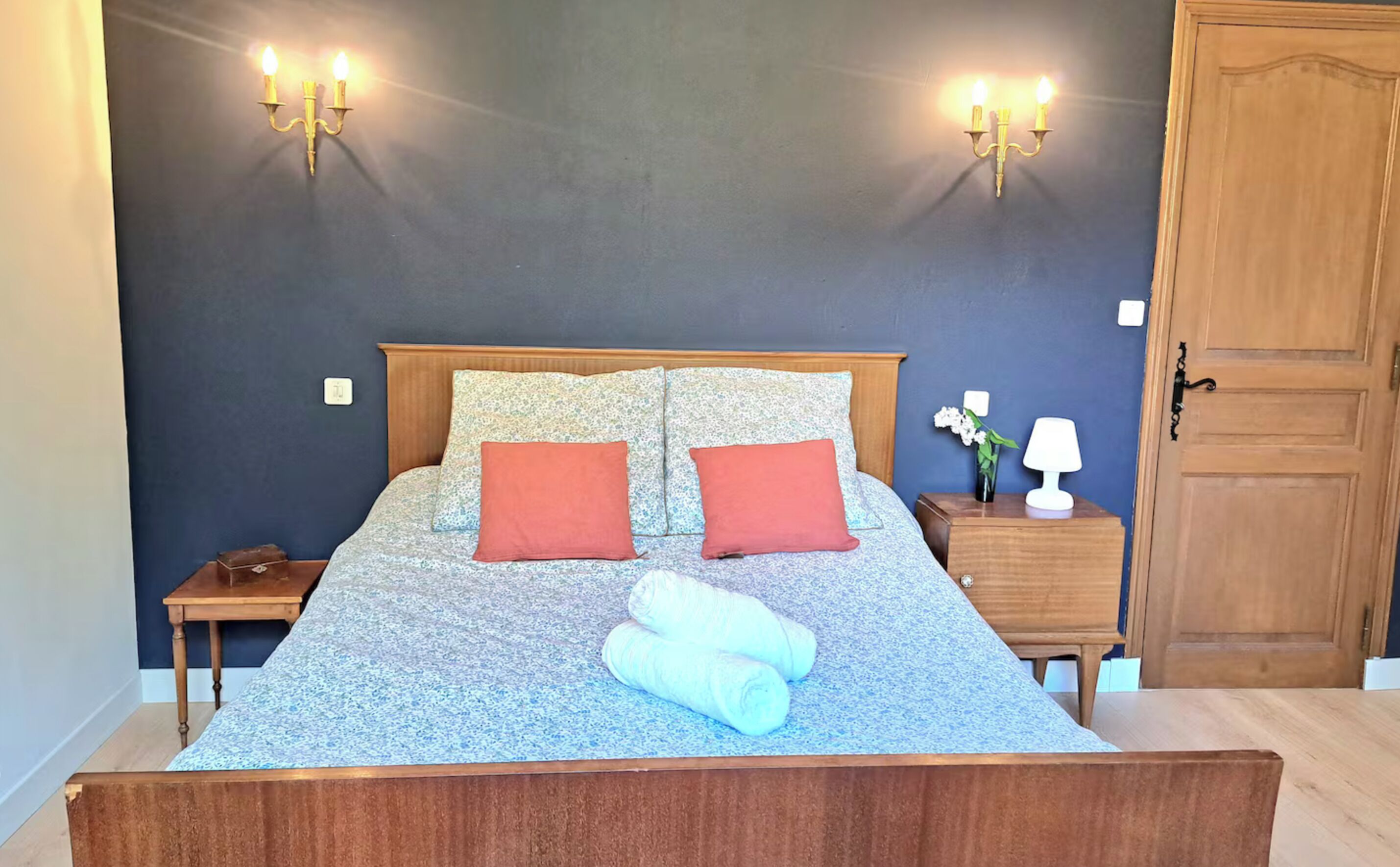 Una cama bien hecha con una colcha floral, dos almohadas rojas y un par de toallas enrolladas. Hay dos mesitas de noche con lámparas y una mesita de noche con un jarrón de flores.