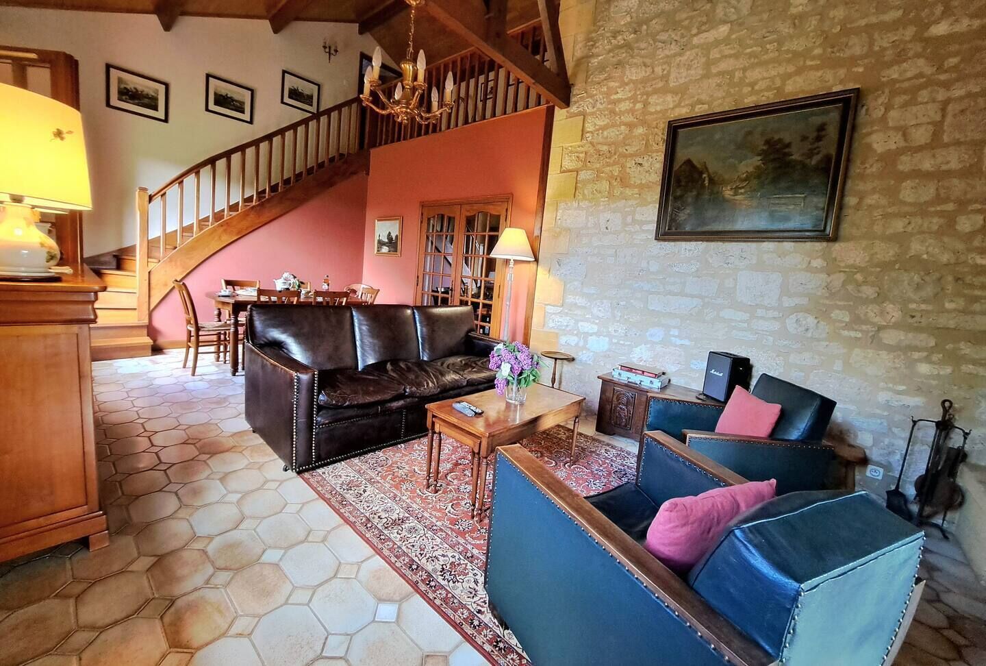 Un salon chaleureux avec un canapé en cuir marron, un fauteuil bleu et une table basse en bois. Un escalier mène à l’étage supérieur, et un tableau est accroché au mur.