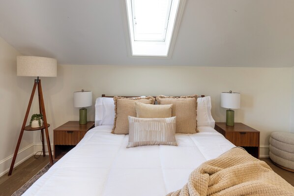 Un dormitorio con tragaluz, una cama con ropa de cama color beige, dos mesitas de noche con lámparas y una planta pequeña en una de ellas.