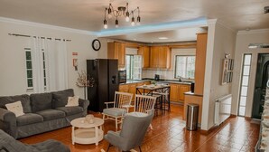 Een moderne woonkamer met een grijze bank, een salontafel en een eethoek met stoelen. In de achtergrond is een keuken met houten kastjes en een raam zichtbaar.