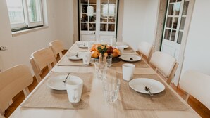 Een eettafel gedekt met borden, kopjes en een schaal fruit, omringd door stoelen in een lichte kamer met ramen.