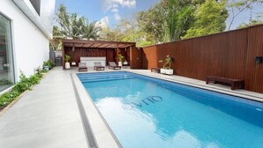 A modern outdoor swimming pool with a wooden deck, lounge chairs, and a pergola. - Luxurious Studio close to USA embassy (Accra)