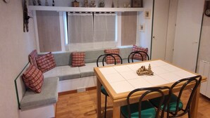 A cozy room with a wooden dining table, four chairs, a bench with cushions, a small shelf with decorative items, and a window with curtains. - MOBIL HOME LOUPIOTS 4/6 PERS. (Besse-et-Saint-Anastaise)