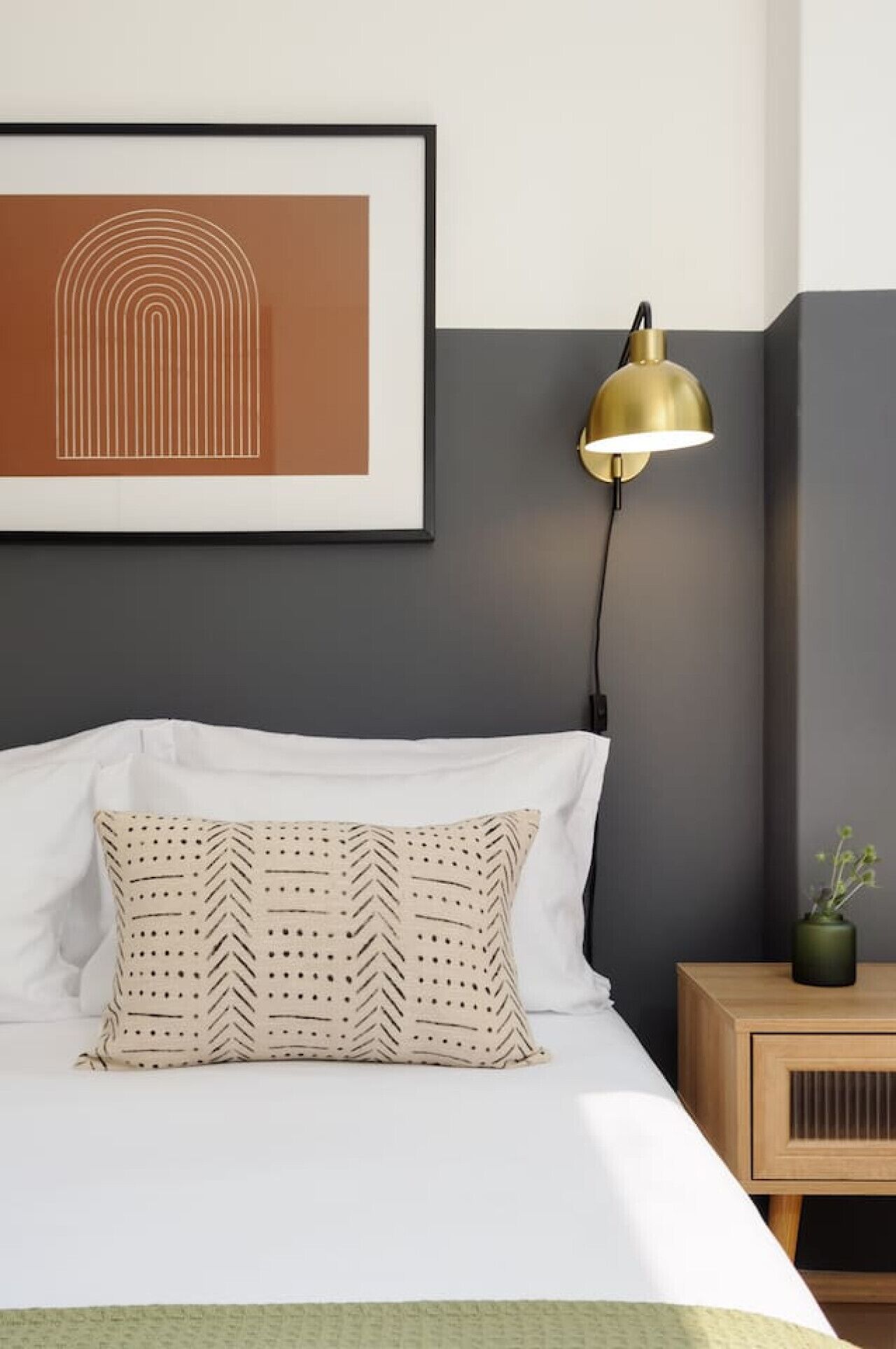 A neatly made bed with white linens and patterned pillows, a gold desk lamp, a framed artwork on the wall, and a small wooden nightstand with a plant.