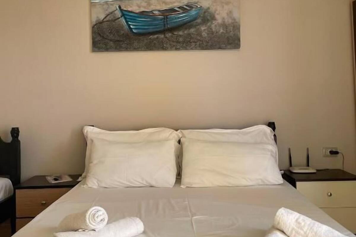 A neatly made bed with white linens and towels, a painting of a boat on a beach, and a simple nightstand with a lamp.