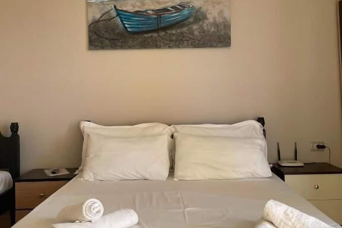 A neatly made bed with white linens and towels, a painting of a boat on a beach, and a simple nightstand with a lamp.