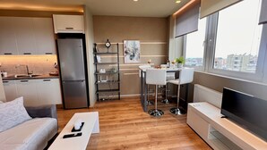 A modern kitchen with a refrigerator, a dining area with a table and chairs, a TV, and a large window with a city view.