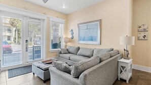 A living room with a grey sectional sofa, a small ottoman, a side table with a lamp, and a framed seahorse artwork on the wall.