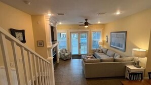 A spacious living room with a large sofa, a coffee table, and a fireplace. There is a staircase leading to another level, and a ceiling fan is visible.