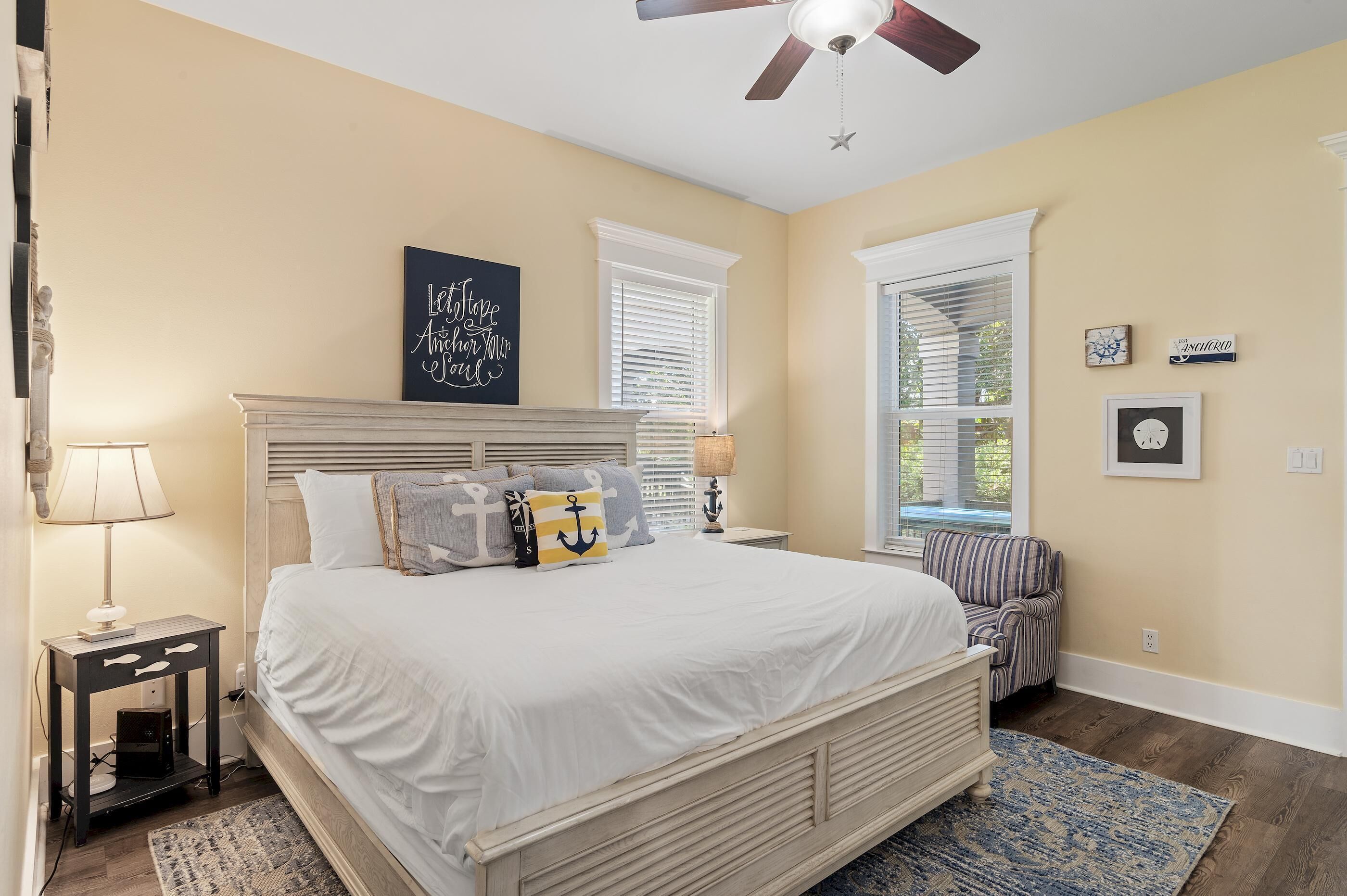 A bedroom with a large bed, a nightstand, a lamp, a ceiling fan, a window with blinds, and a small chair.