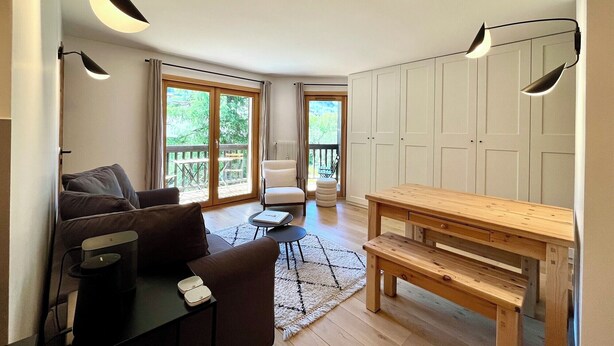 Een moderne woonkamer met een bank, een houten zitbank en een eettafel. Er zijn twee deuren die leiden naar balkons met buitenmeubilair.