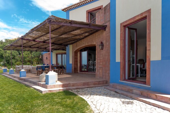 A covered patio area with outdoor furniture, a stone pathway, and a building with blue and beige walls. - Beachfront Recluse by Cadenzalux (Sintra)