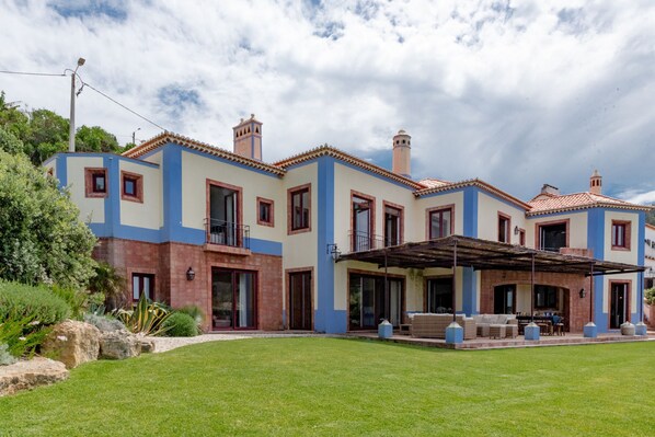 A two-story house with a patio area, large windows, and a well-maintained lawn. - Beachfront Recluse by Cadenzalux (Sintra)