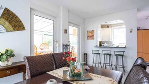A dining area with a table set for two, a vase with flowers, and a view of the kitchen in the background.