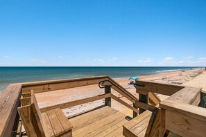 Terraza de madera con vista a una playa y al océano, donde se ve un paraguas a lo lejos.