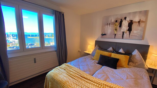 A hotel room with a bed, a painting on the wall, a window with a view of the sea, and a lamp on the side table.