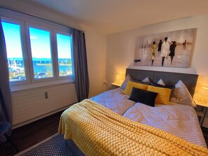 Ein Hotelzimmer mit Bett, einem Gemälde an der Wand, einem Fenster mit Meerblick und einer Lampe auf dem Nachttisch.