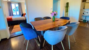 A modern dining area with a wooden table and blue chairs, a small vase with flowers, and a view of a bed and a window with curtains.