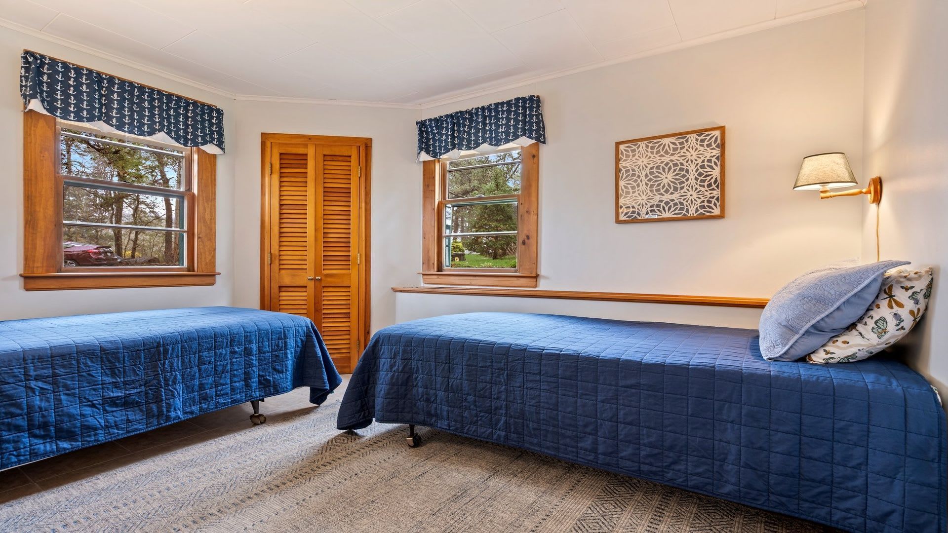 A bedroom with two beds, a wooden door, windows with curtains, a wall-mounted lamp, and a decorative piece.