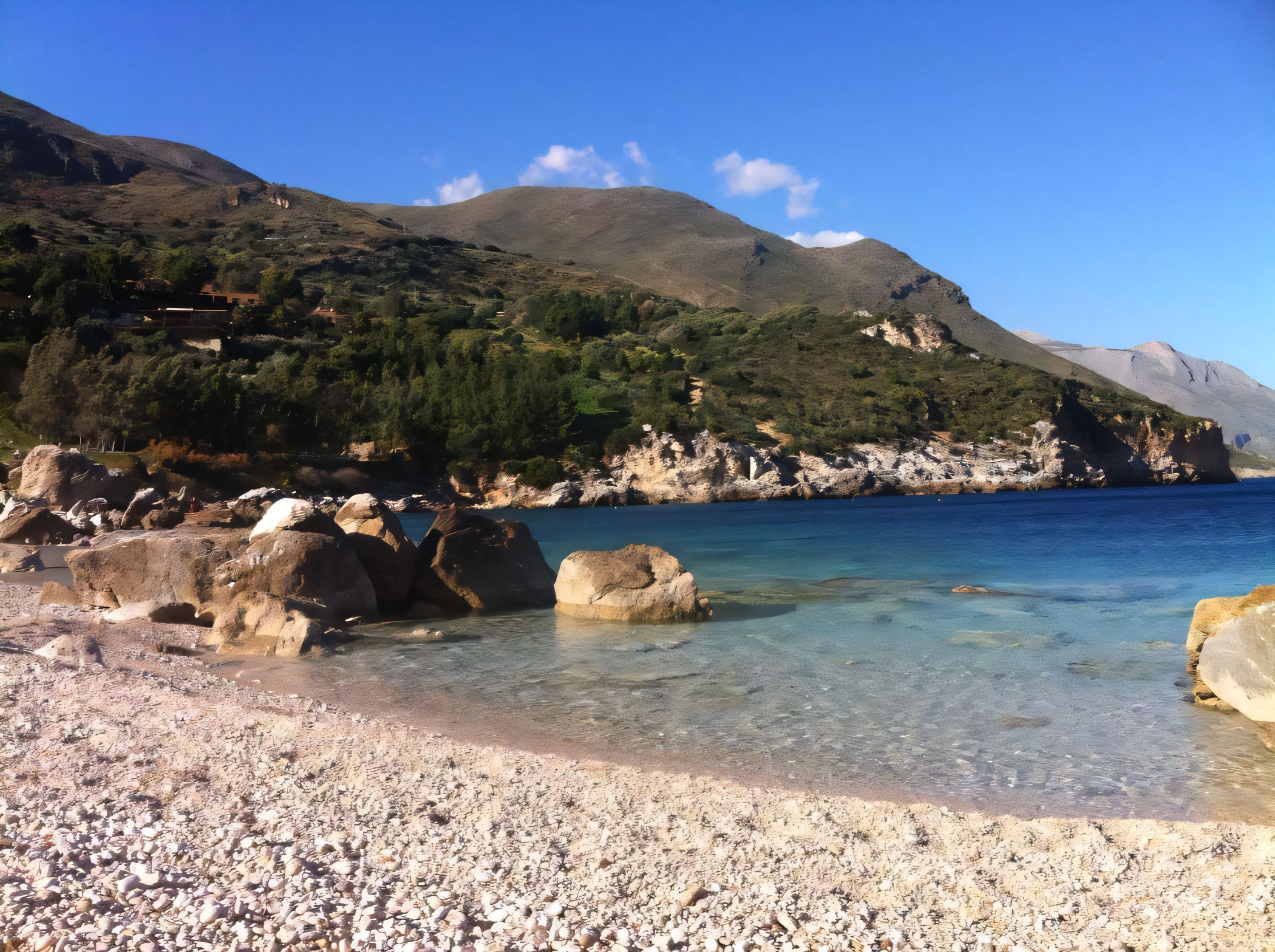 A pebble beach with clear water, large rocks, and a mountainous backdrop.