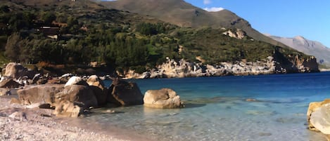 A pebble beach with clear water, large rocks, and a mountainous backdrop.