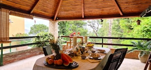 An outdoor dining area with a wooden ceiling, a table set for two with fruit and drinks, and a view of greenery and trees. - Villa Dream House Scopello (Castellammare del Golfo)