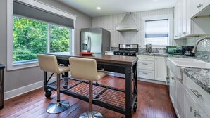 A modern kitchen with a central island, two bar stools, a stainless steel refrigerator, a gas stove, and a large window with a view of greenery.