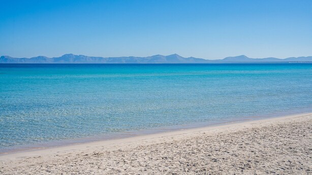 Une plage bordée d’une eau turquoise et limpide, avec des montagnes lointaines se découpant sur un ciel bleu azur.