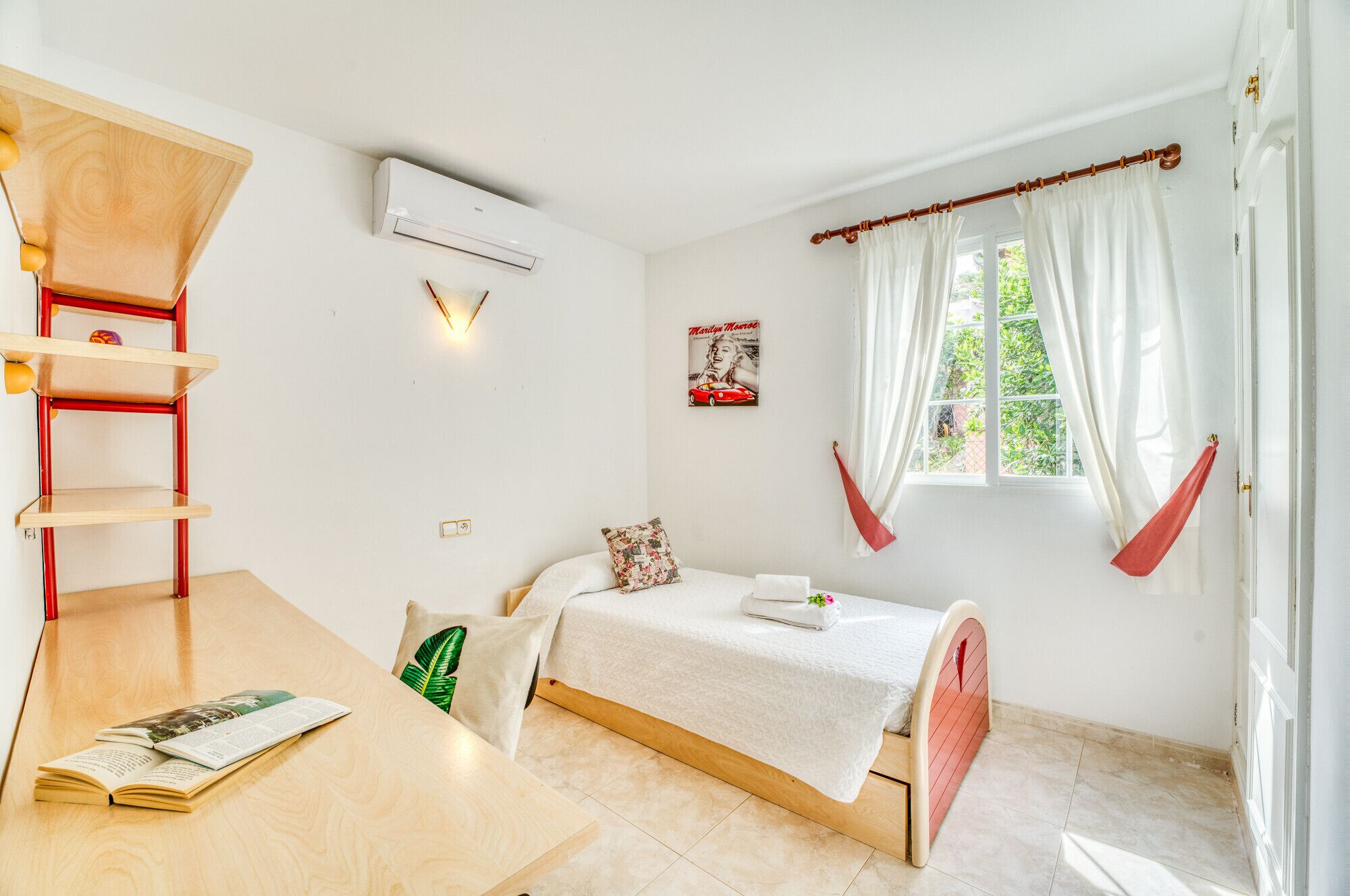 A bedroom with a bed, a wooden shelf unit, a wall-mounted air conditioner, a window with curtains, and a poster on the wall.