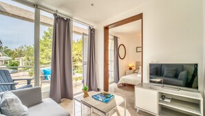 Une chambre d’hôtel moderne avec un canapé, une télévision, une petite table avec un magazine et un balcon donnant sur une piscine.