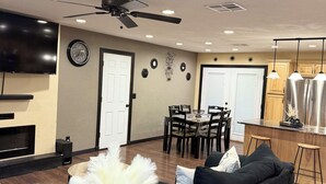 A modern living room with a black sofa, a small table with a vase, a dining area with a table and chairs, and a kitchen area with a refrigerator and pendant lights.