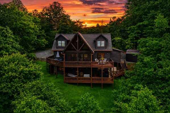 A two-story log cabin with a deck and a covered porch, surrounded by dense forest. - Sunrock Chalet: Discover Modern Luxe Vibes • Hot-Tub • Pool Table • Epic Views (Blue Ridge)