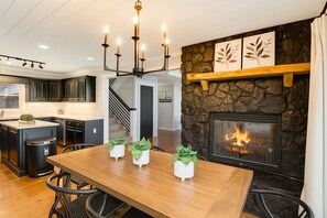 A modern kitchen with dark cabinets, a wooden dining table with potted plants, and a stone fireplace with a visible fire. - Sunrock Chalet: Discover Modern Luxe Vibes • Hot-Tub • Pool Table • Epic Views (Blue Ridge)