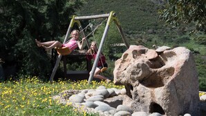 Une balançoire en bois avec deux enfants dessus, entourée d’un paysage naturel composé de rochers et de fleurs jaunes.