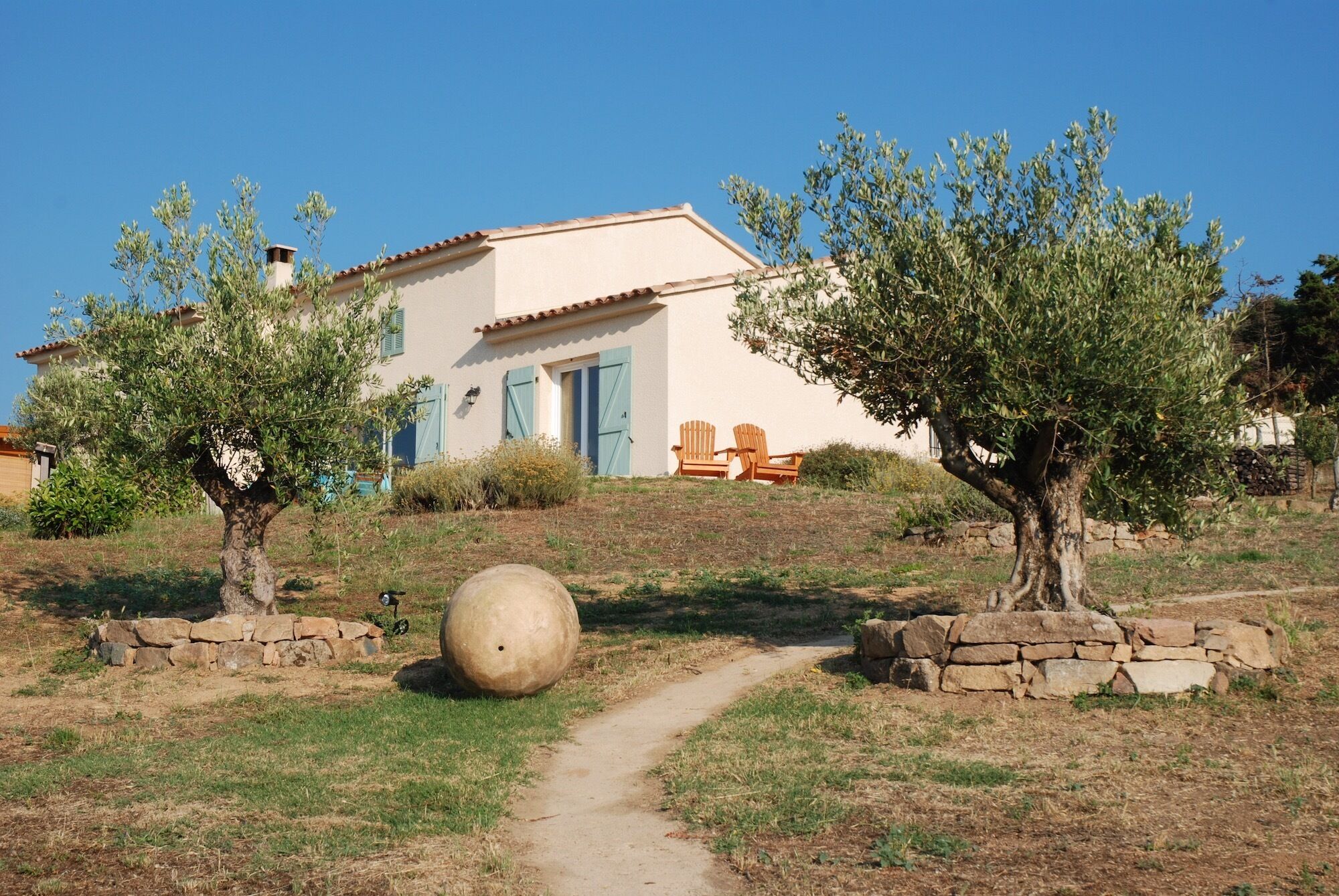 A house with a pathway leading to a door, surrounded by olive trees and a large stone.