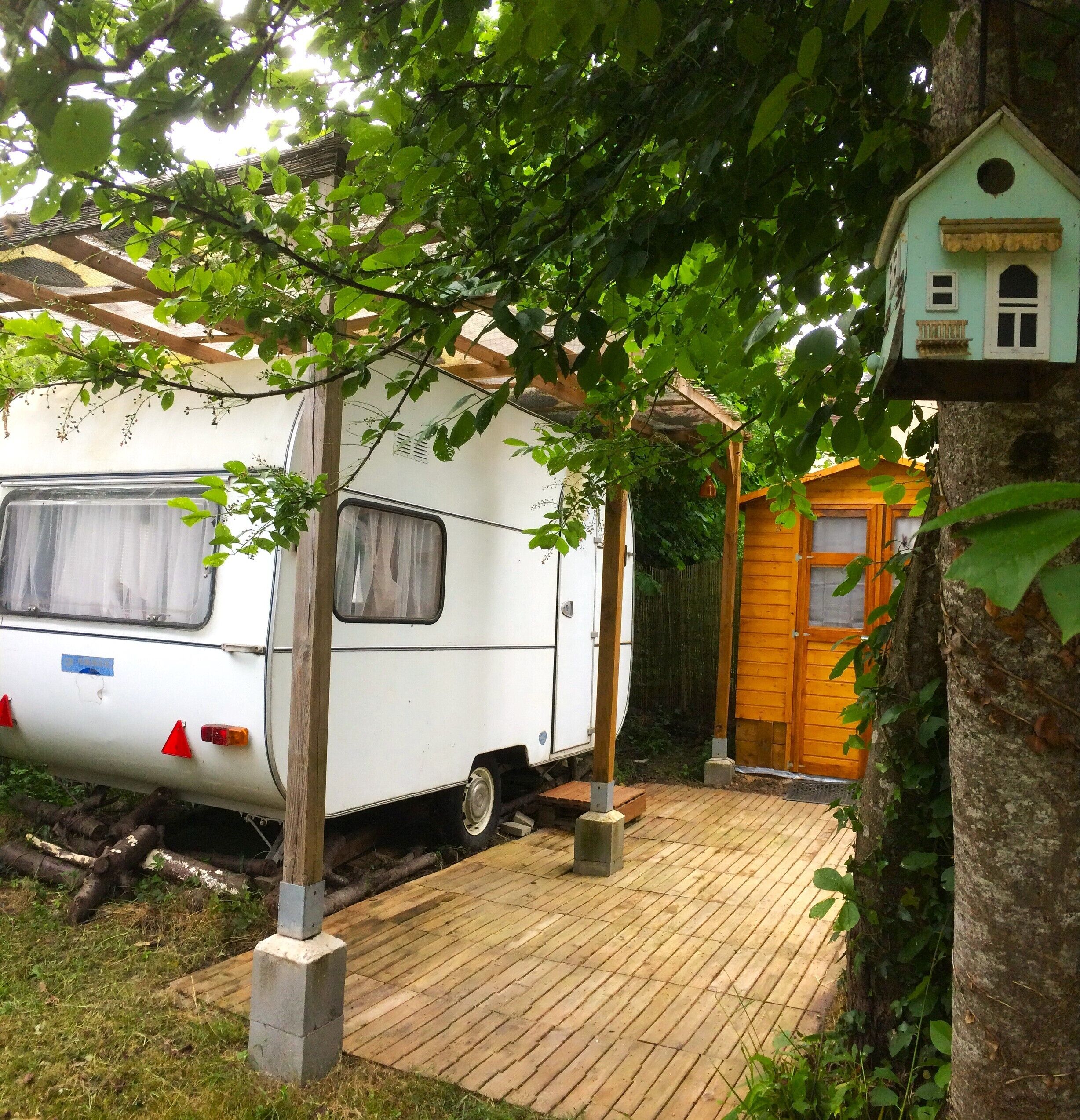 Un camping-car blanc est garé sous un arbre, à côté d’une terrasse en bois et d’une petite construction en bois dotée d’une porte.