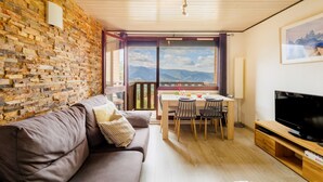 Ein modernes Wohnzimmer mit Backsteinwand, Holzboden, einem großen Fenster mit Bergblick, einem braunen Sofa, einem Esstisch mit Stühlen und einem Flachbildfernseher.
