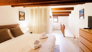 Ein Schlafzimmer mit einem Bett, einem Holz-Kopfende, weißen Wänden und einer Holzbalkendecke. Es gibt eine Holzkommode und einen Fernseher auf einem Schrank.