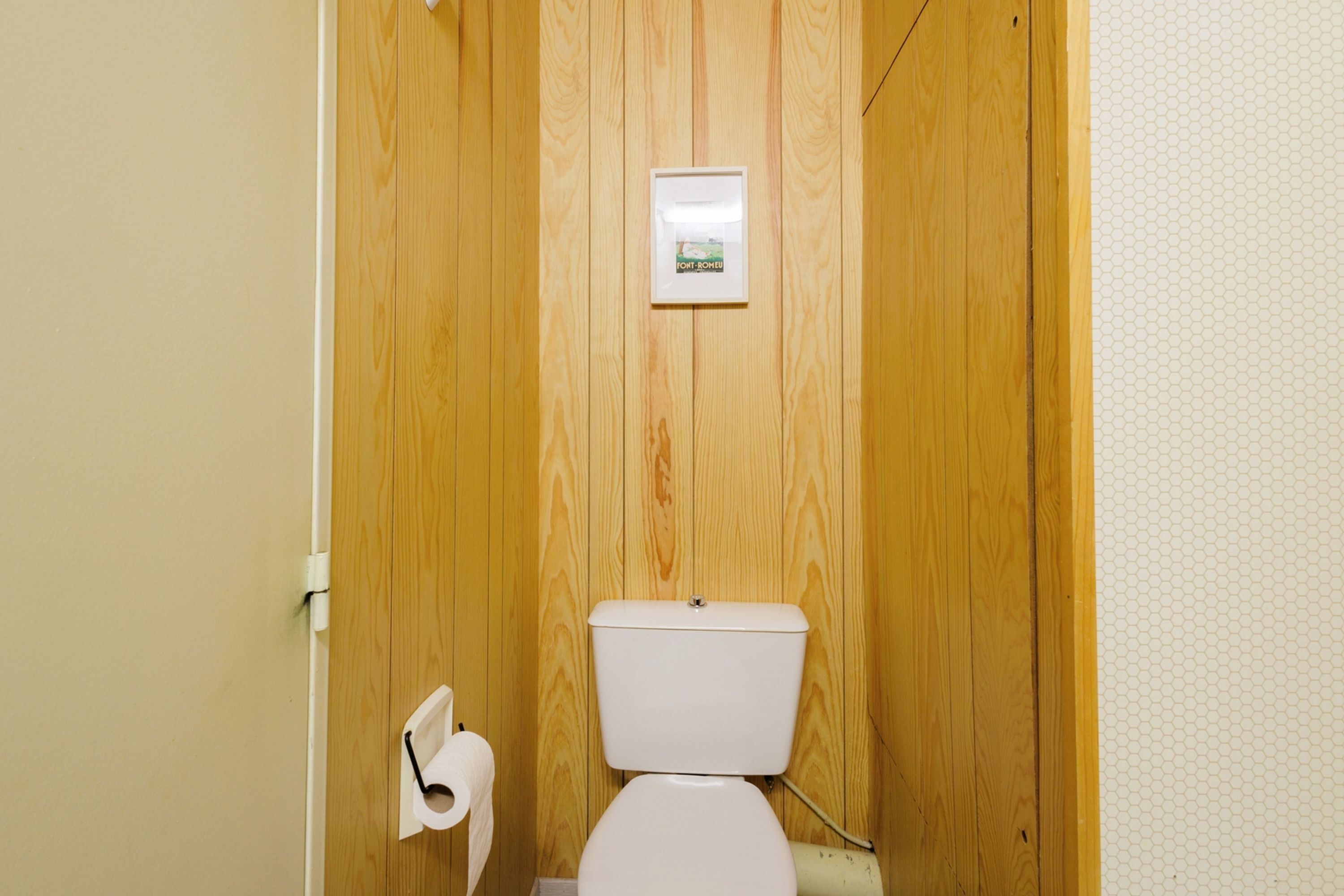 Une petite salle de bain aux murs lambrissés, équipée de toilettes, d’un porte-rouleau de papier toilette et ornée d’un tableau.