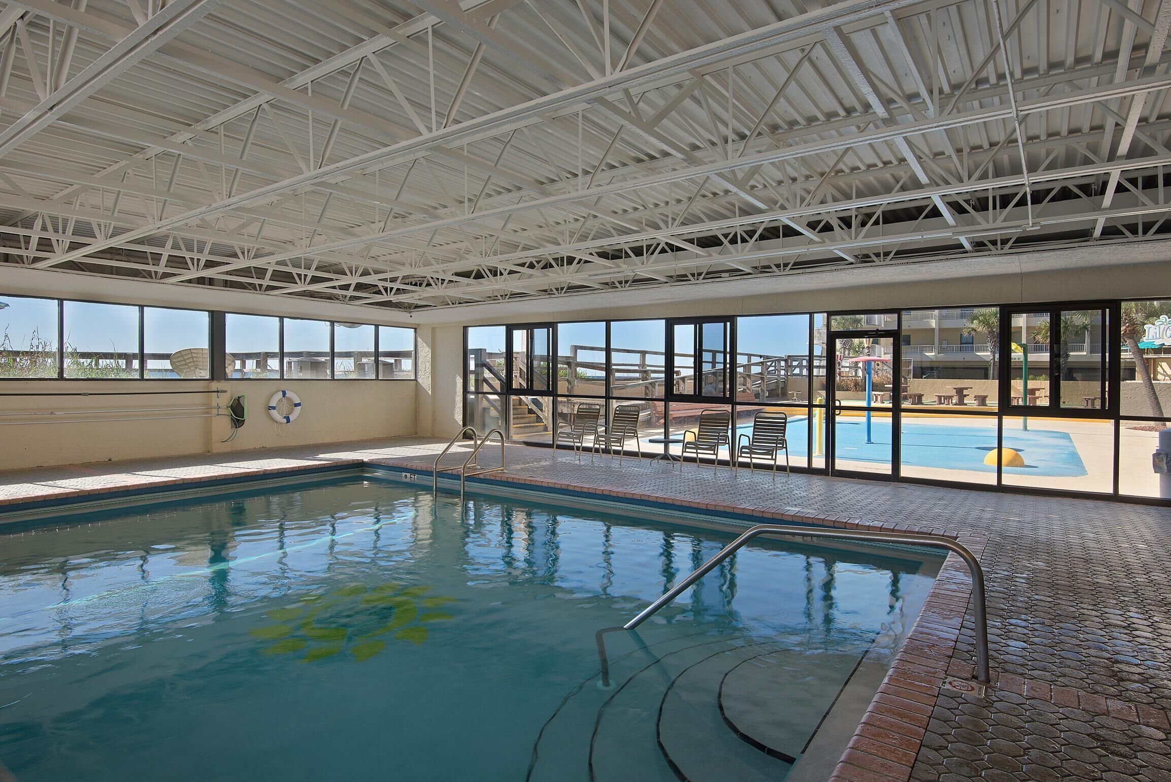 Une piscine intérieure avec des parois en verre transparent, un sol carrelé et une structure de toit en métal.