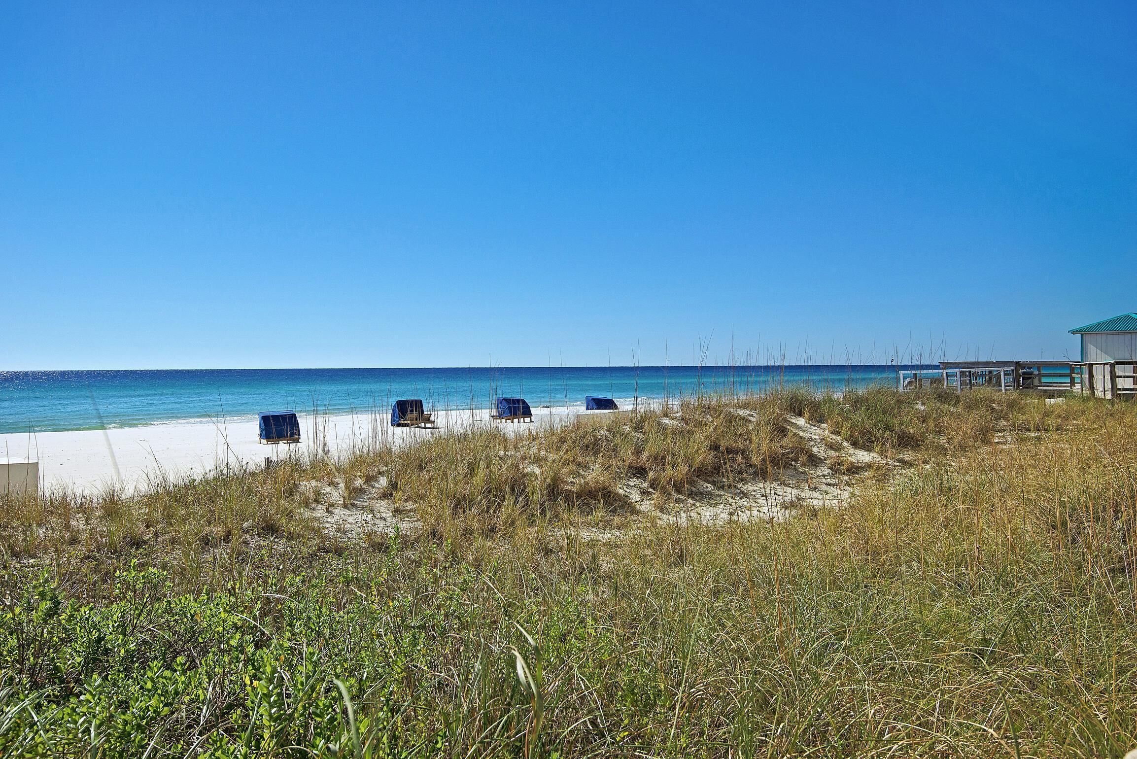 Une plage de sable blanc, avec une eau d’un bleu limpide et quelques chaises de plage bleues. On aperçoit, au loin, un bâtiment surmonté d’un toit vert.
