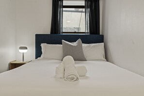Una cama bien hecha con almohadas, una toalla enrollada y una lámpara de noche en una habitación con ventana y cortinas.