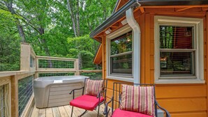 Une terrasse en bois avec un jacuzzi, deux fauteuils à bascule avec des coussins rouges et une fenêtre avec des stores.