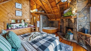 A cozy cabin interior with a wooden ceiling, stone fireplace, and a large bed with a checkered blanket. - Hot Tub! Cute Studio Cabin! Views! Private! Relax! (Sevierville)