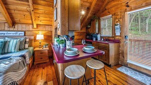 A wooden cabin interior with a kitchen area, a dining table with stools, a bed with a sign, and a window with blinds. - Hot Tub! Cute Studio Cabin! Views! Private! Relax! (Sevierville)