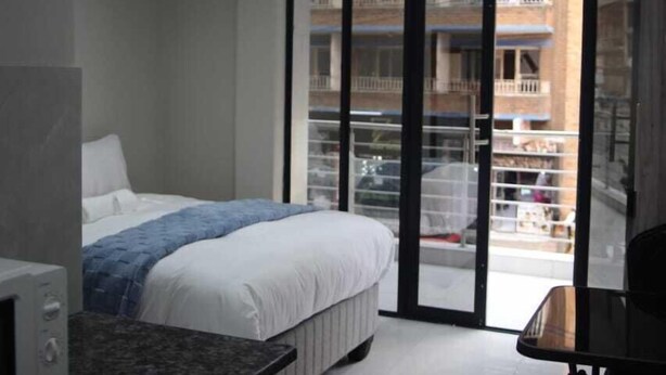 Una habitación de hotel moderna con una cama, una kitchenette pequeña y un balcón al que se accede por una puerta corrediza de vidrio.
