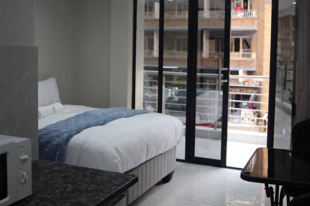 A modern hotel room with a bed, a small kitchenette, and a sliding glass door leading to a balcony.