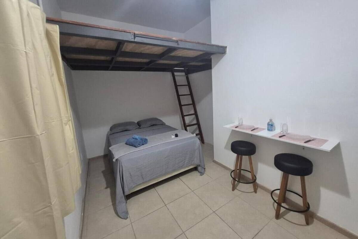 Una habitación pequeña y funcional con una cama individual superpuesta, un escritorio y dos taburetes, además de una escalera.